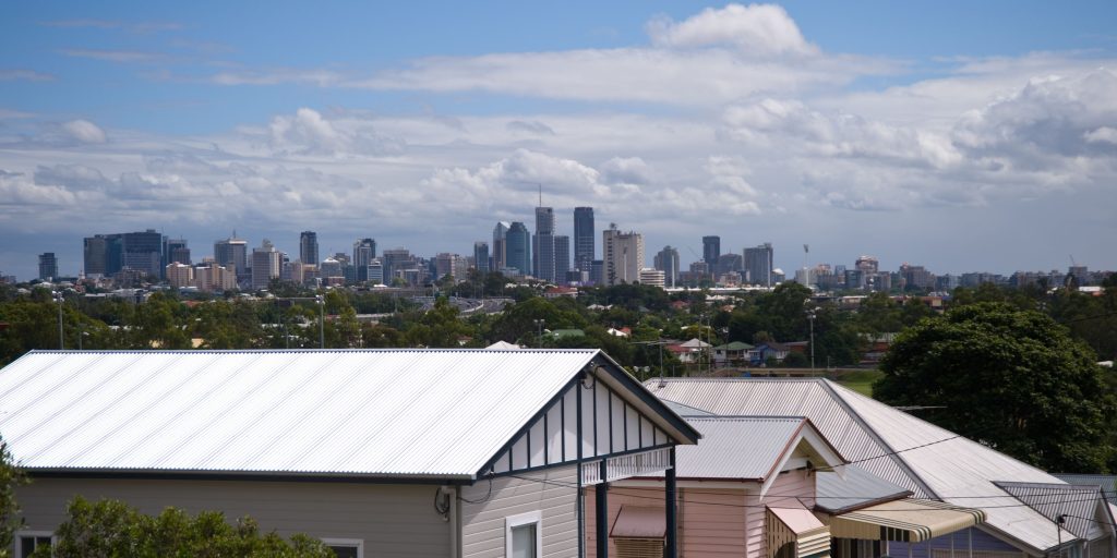 A white and pink Queenslander on a hill in a Brisbane suburban street, with a city skyline in the background.